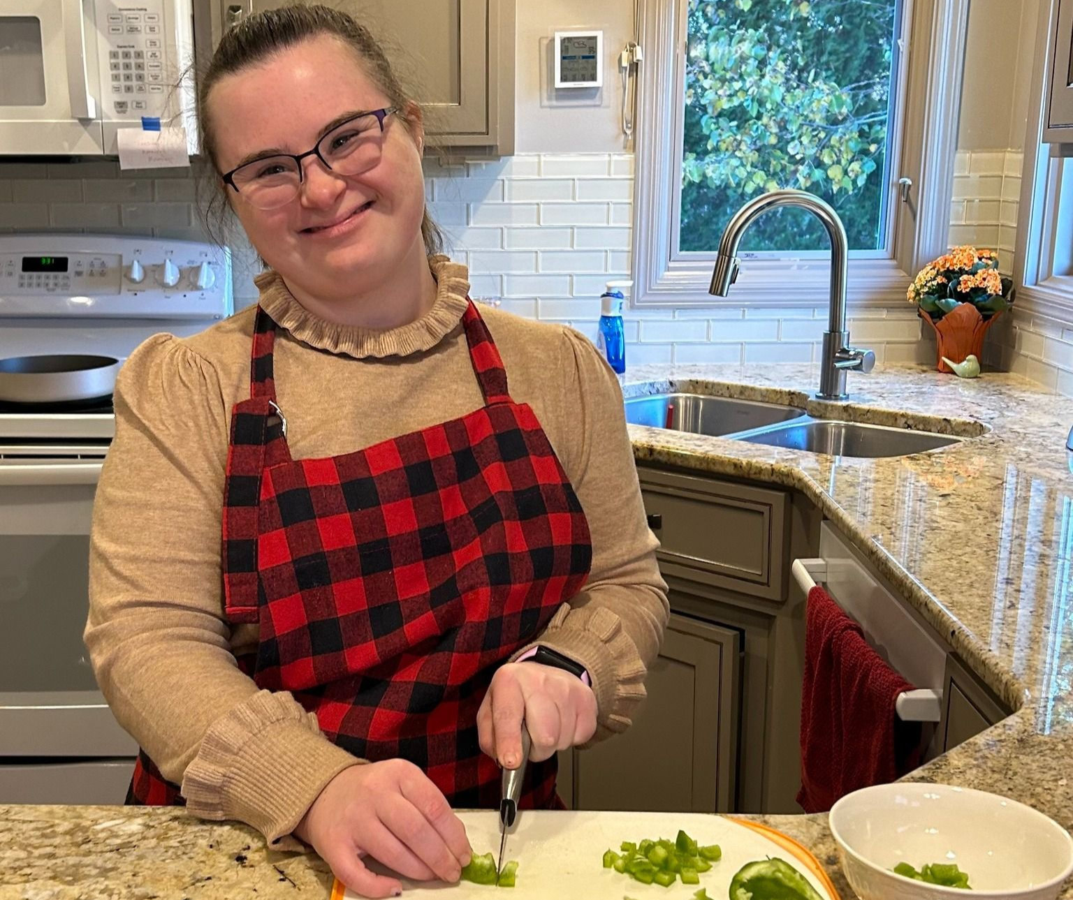 Woman with glasses and apron, chopping vegetables in a kitchen, smiling.