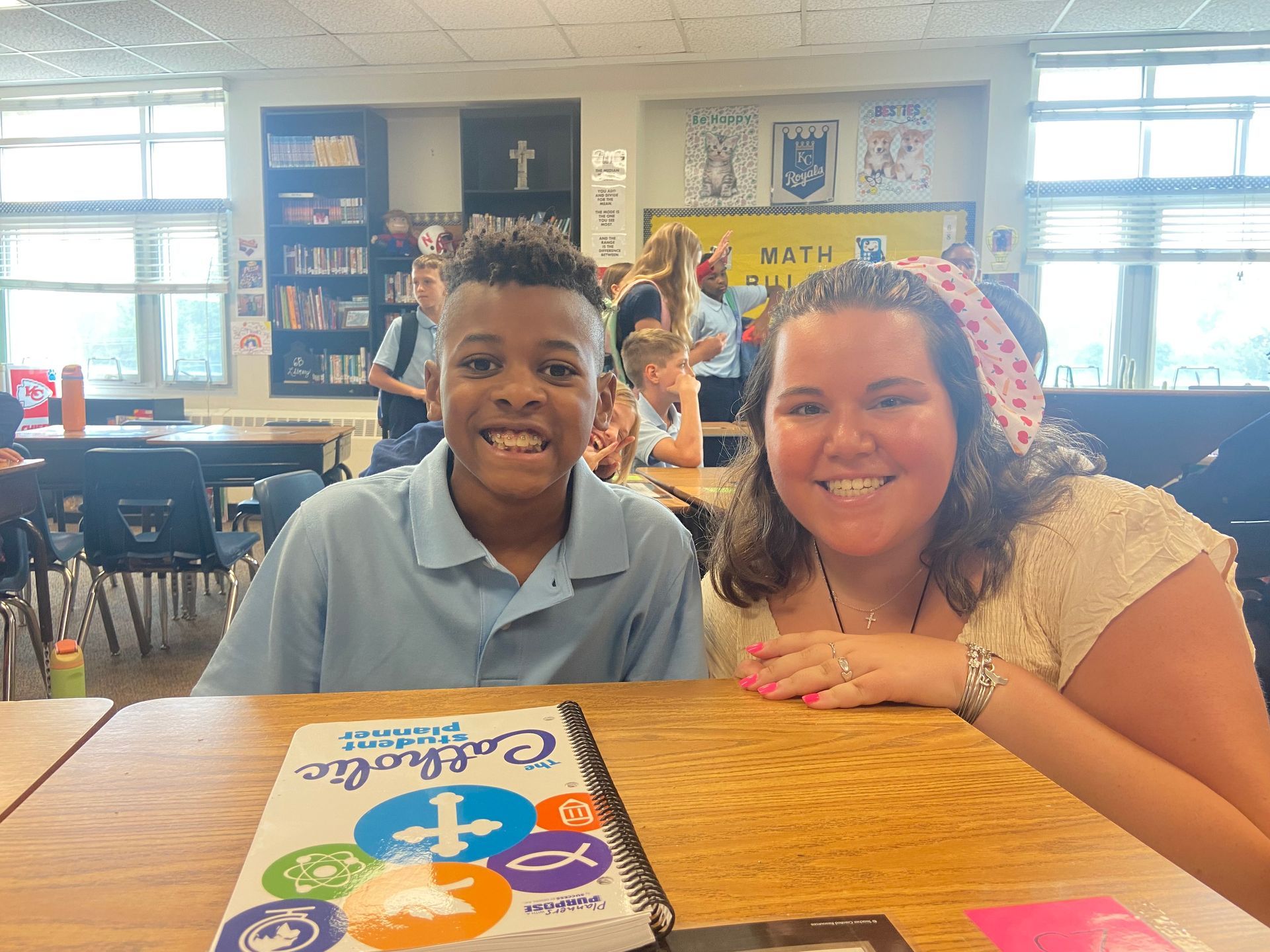 A smiling person and child seated at a desk, book in front. School interior.