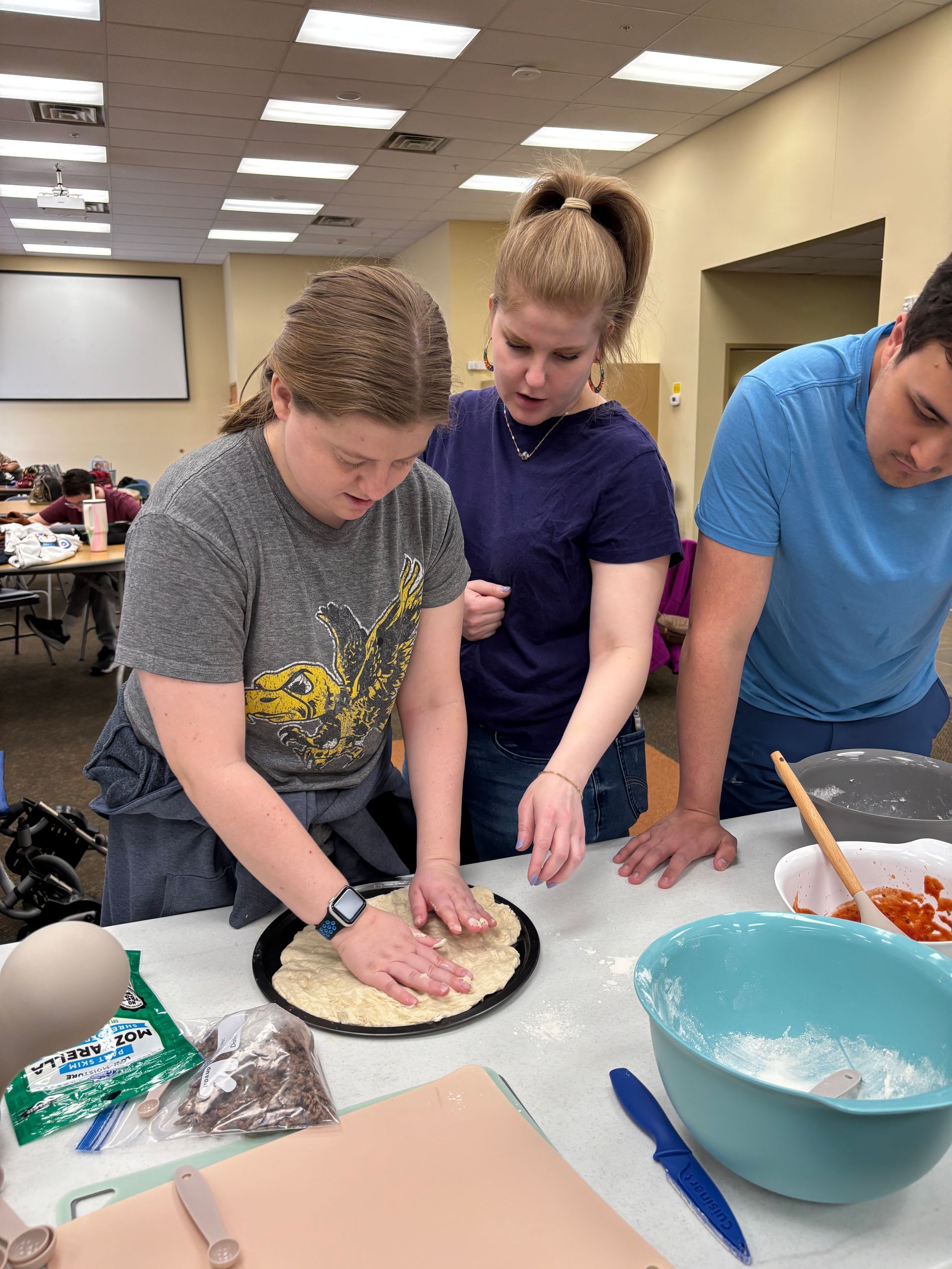 Three people making pizza together at a table in a room. One person presses dough, another guides.