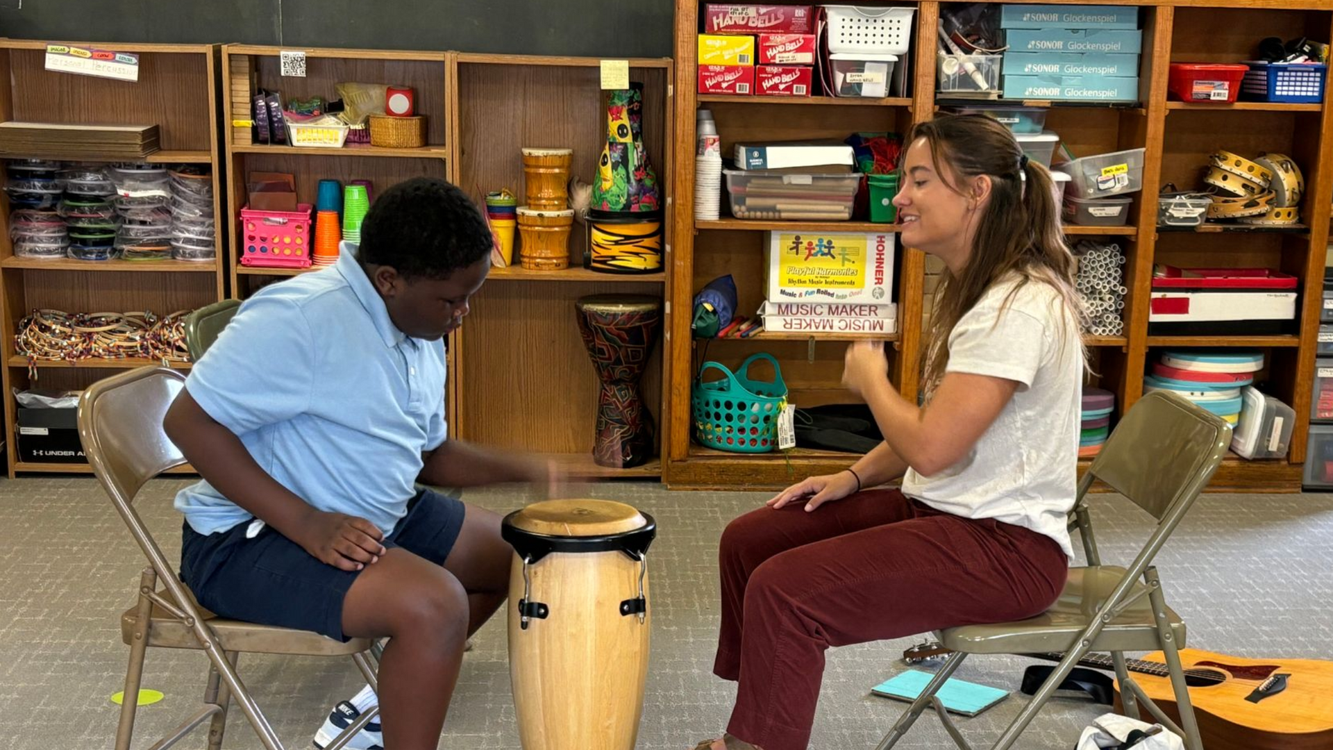 Person playing a drum with a teacher in a music room.