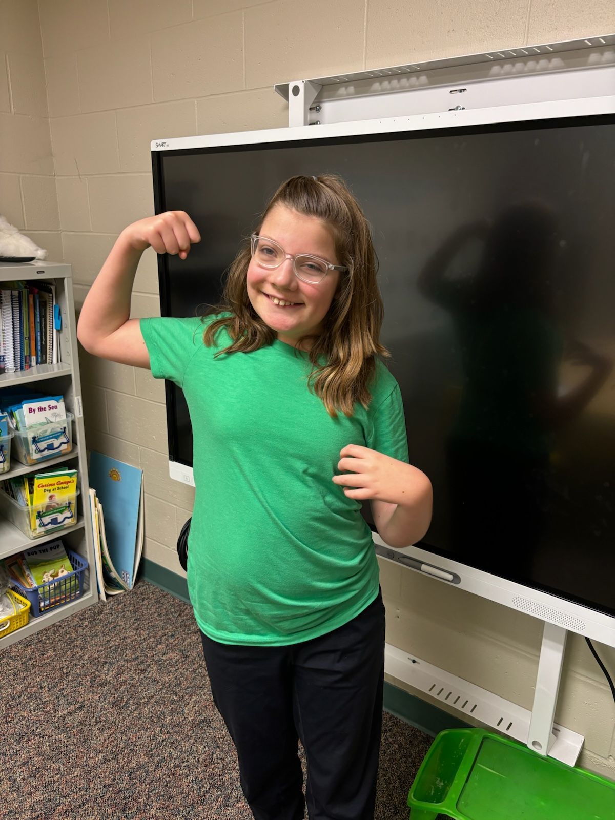 Girl in green shirt flexing arm, standing in front of a smartboard.