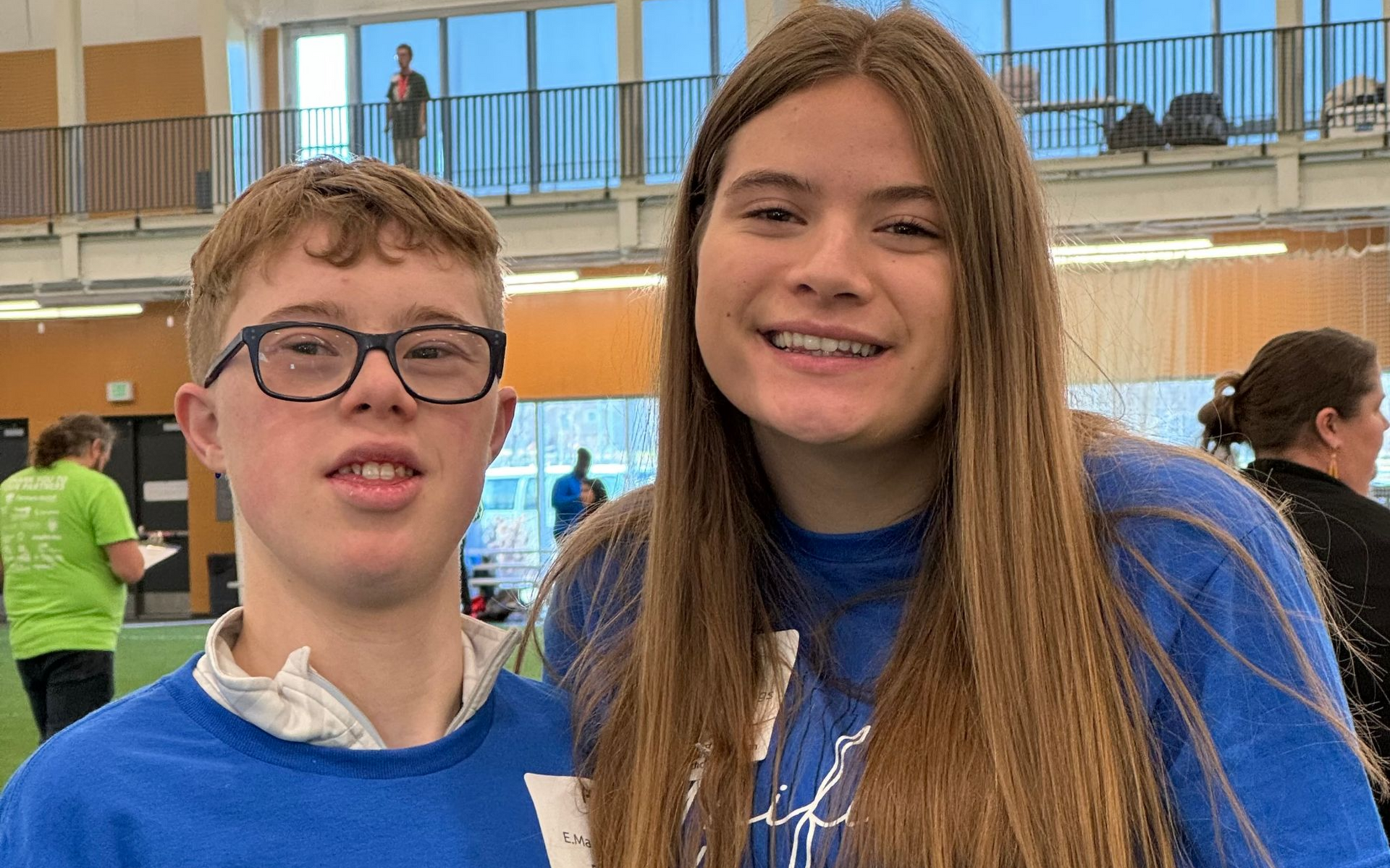 Two people smiling inside a building. One has glasses, the other long brown hair. Both wear blue shirts.