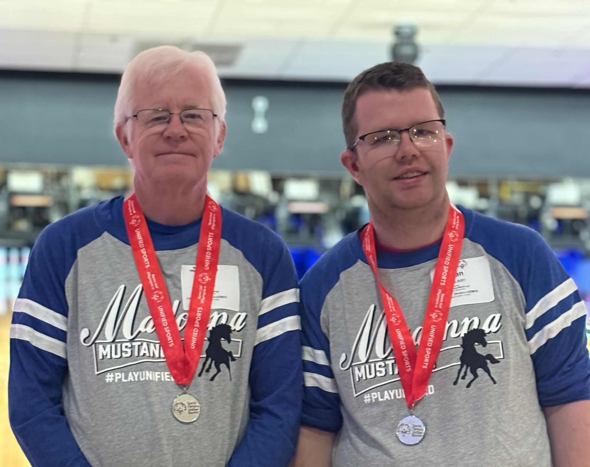 Two men wearing matching bowling shirts and medals pose in a bowling alley.