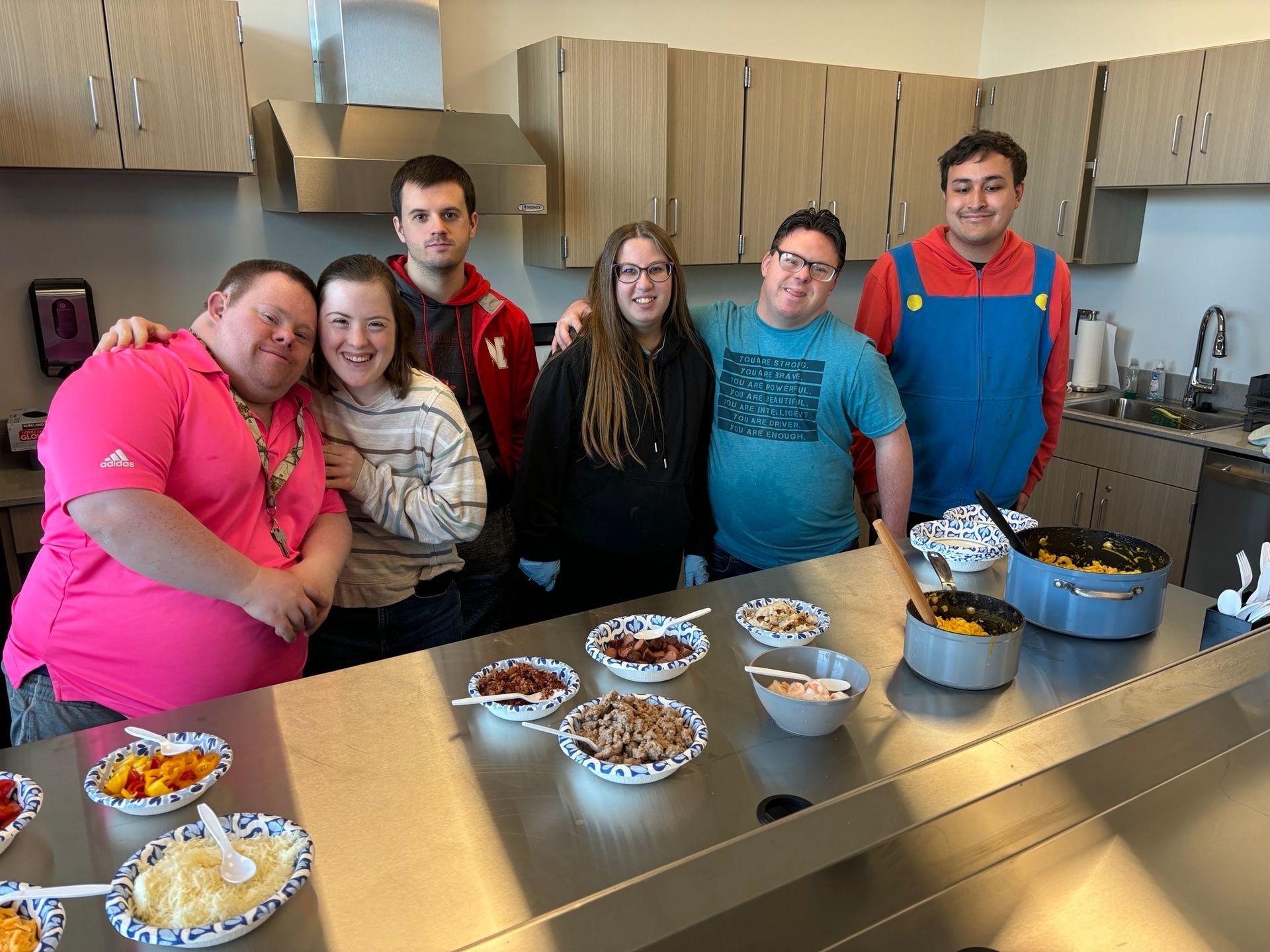 Group of people smiling, standing behind a table with various food dishes in a kitchen.