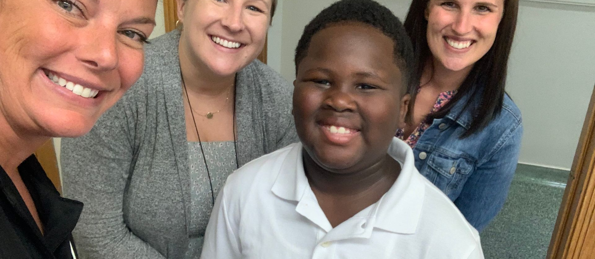 Four smiling people, including a boy in a white shirt, pose together indoors.