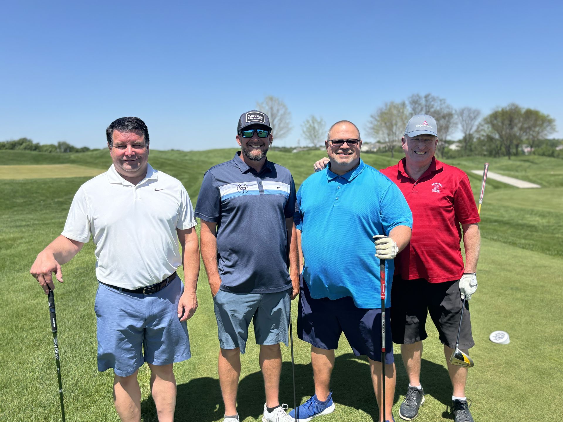 Four men stand on a golf course, smiling with golf clubs. Sunny day, green grass, light clothing.
