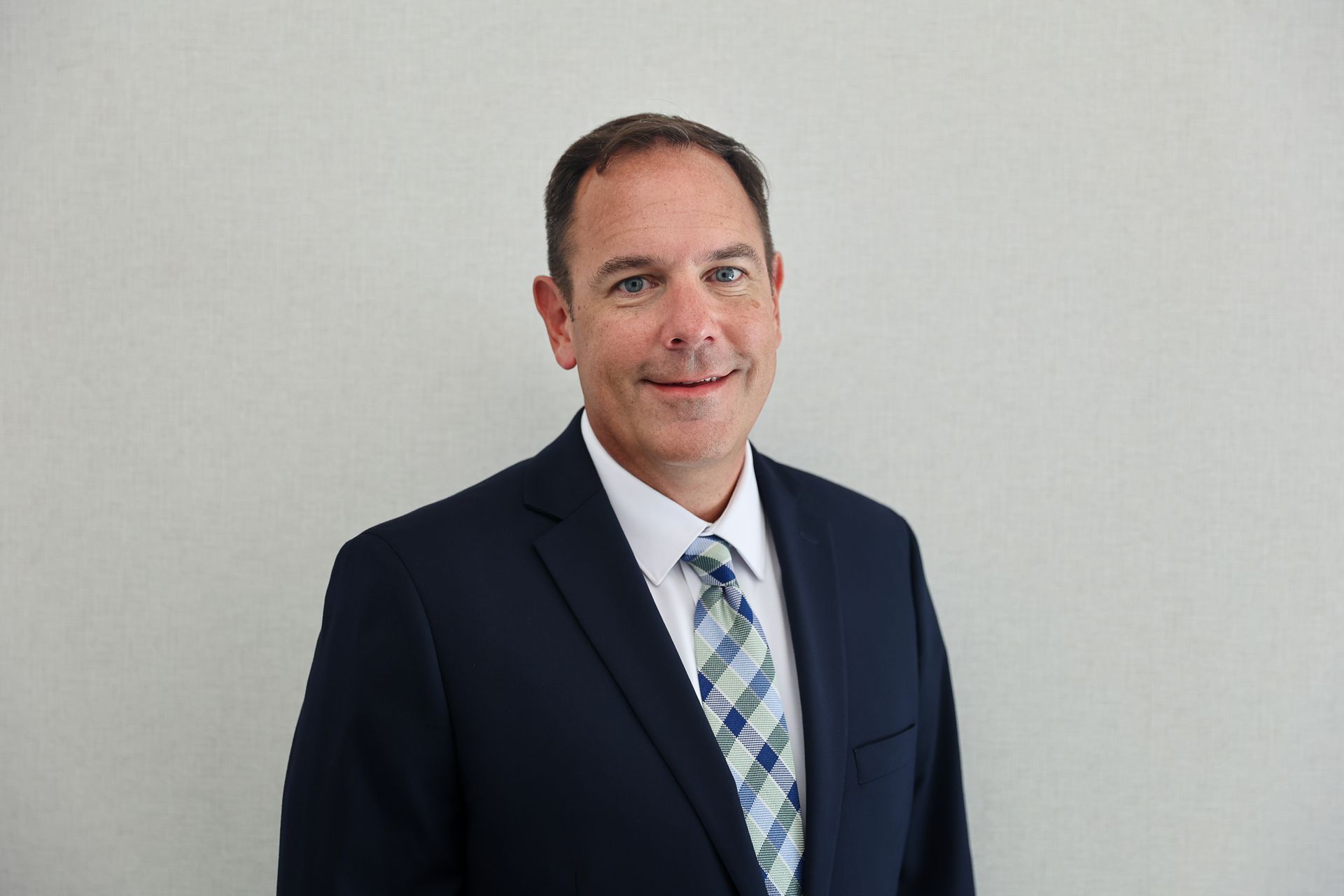Man in suit and tie, smiling, against a light gray wall.