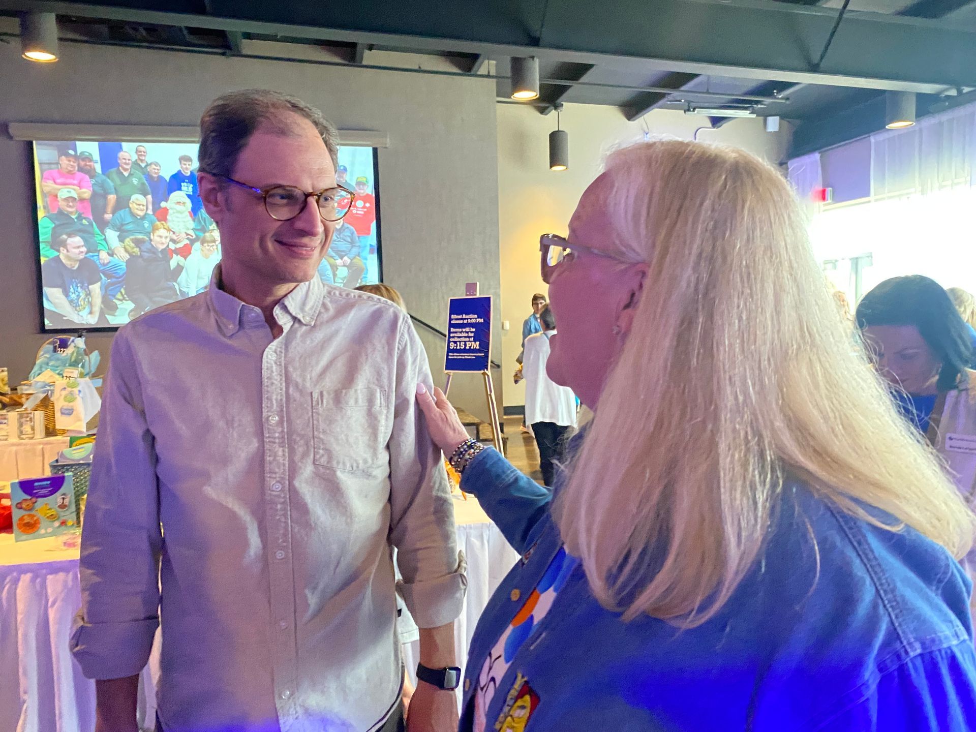Man in glasses and woman with blonde hair, woman touching man's shoulder. Event setting, background with projection screen.
