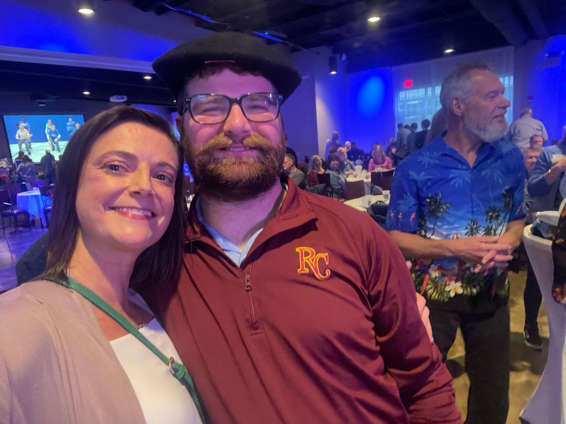 Two people smiling at a formal event, man in maroon shirt and beret, woman in tan jacket. People seated in background.