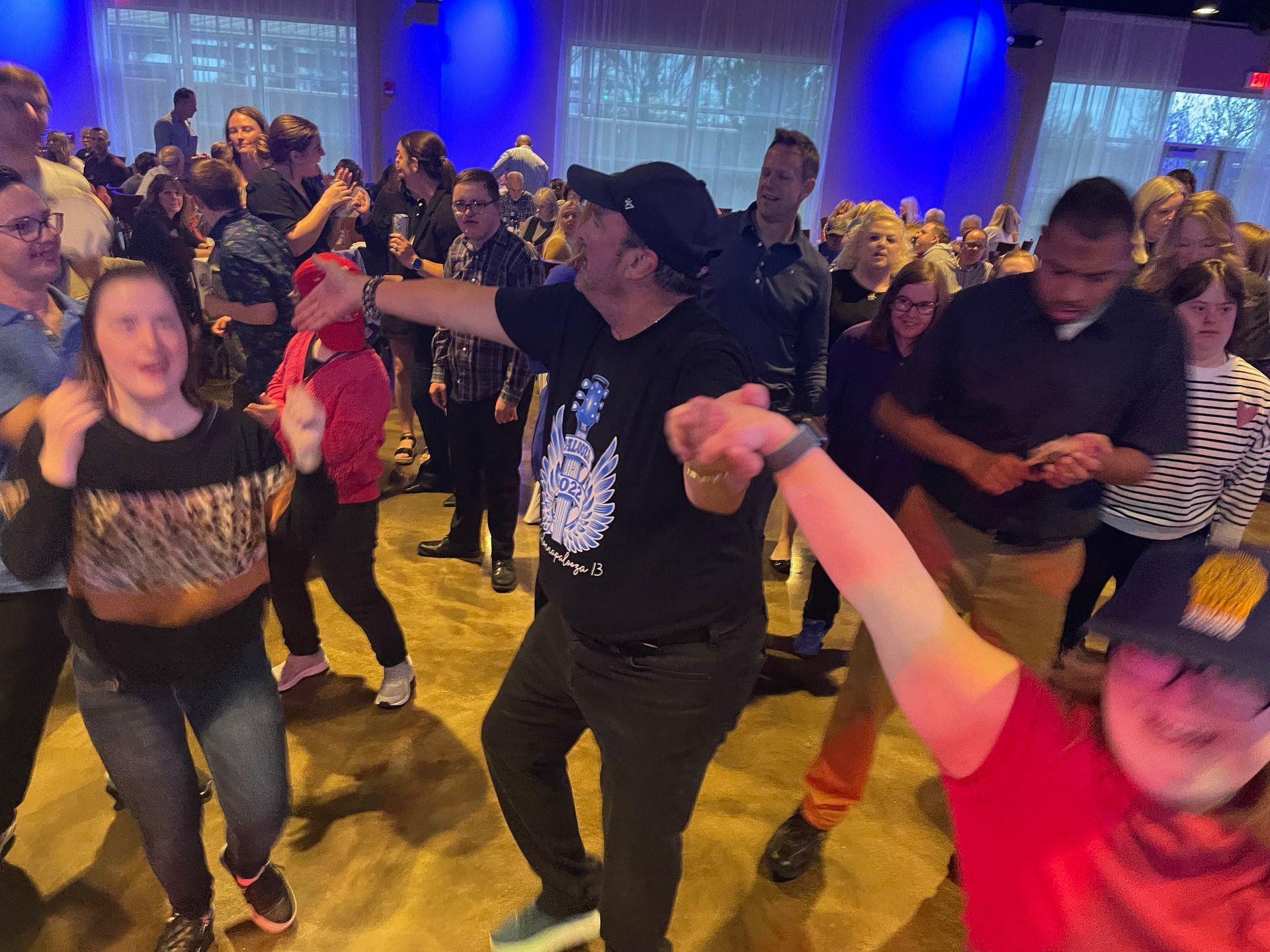 People dancing at an event, arms outstretched. Blue lighting, many individuals in a large room.