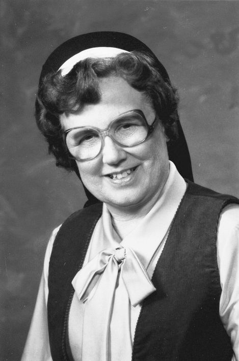 Nun in glasses, smiling, wearing a black veil, white headpiece, and vest over a blouse.