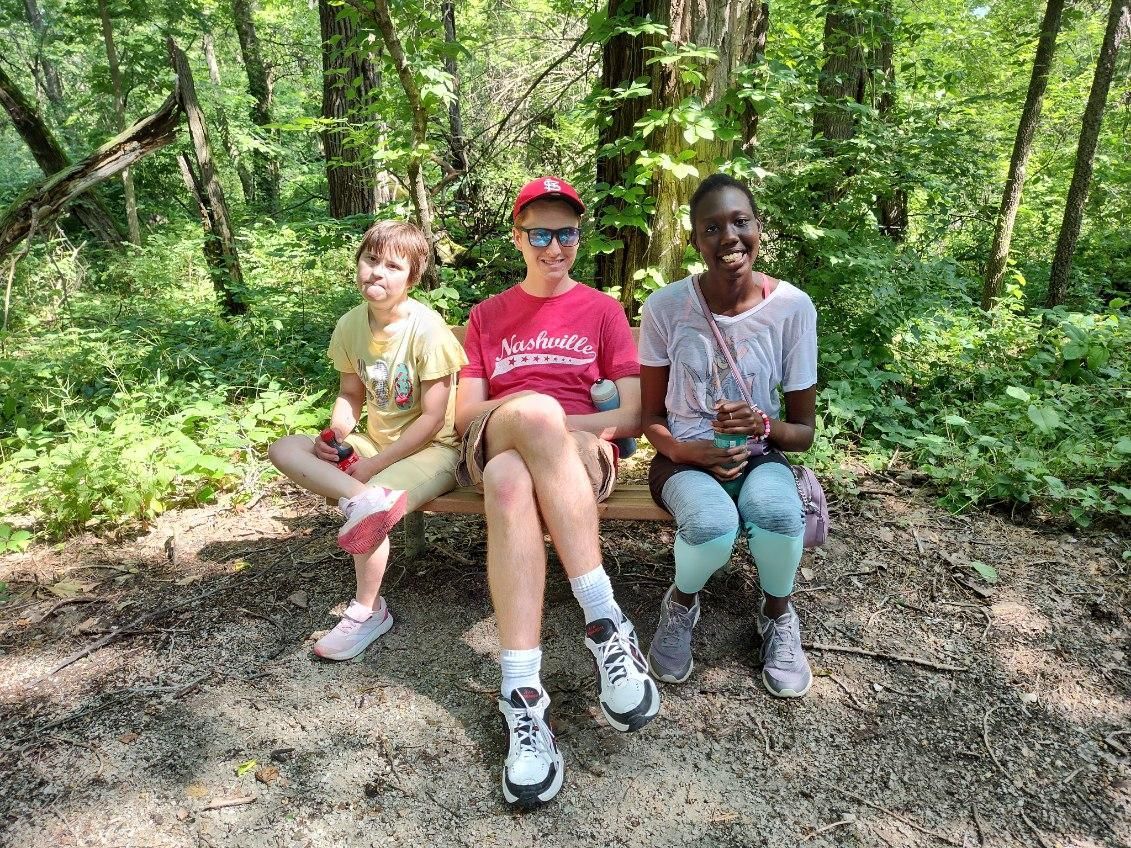 Three people sitting on a bench in a forest. One wears a red shirt, hat, and sunglasses. The others are smiling.
