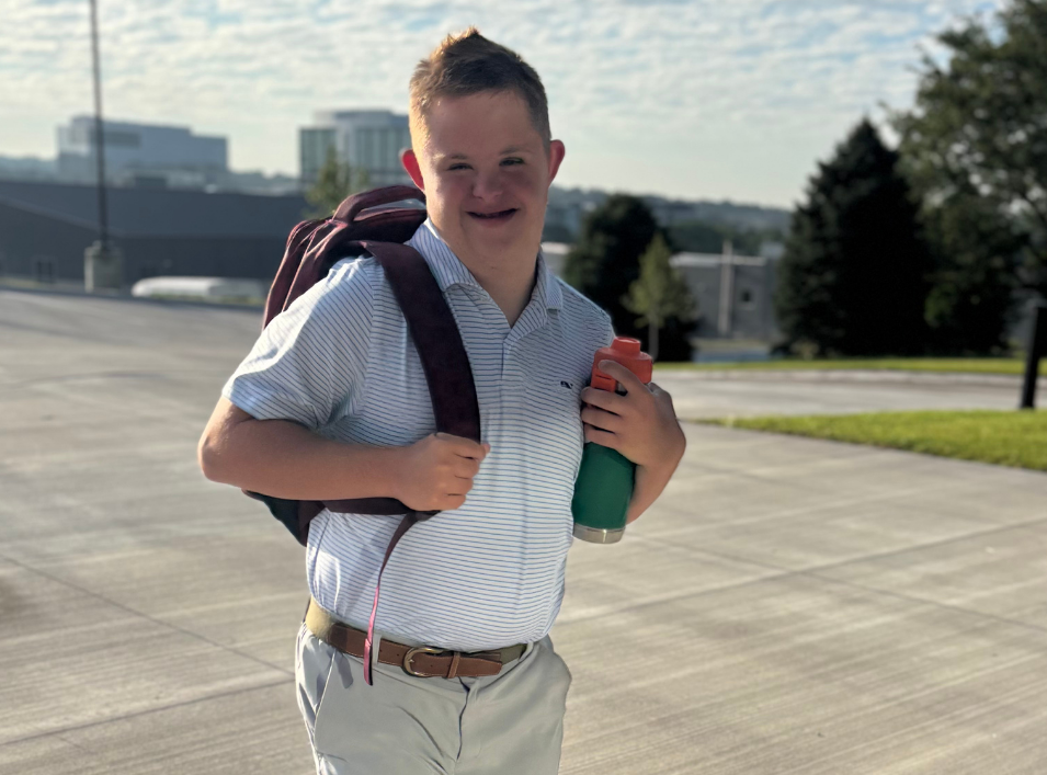 A young person with Down syndrome smiles, wearing a backpack, holding a water bottle, standing outside.