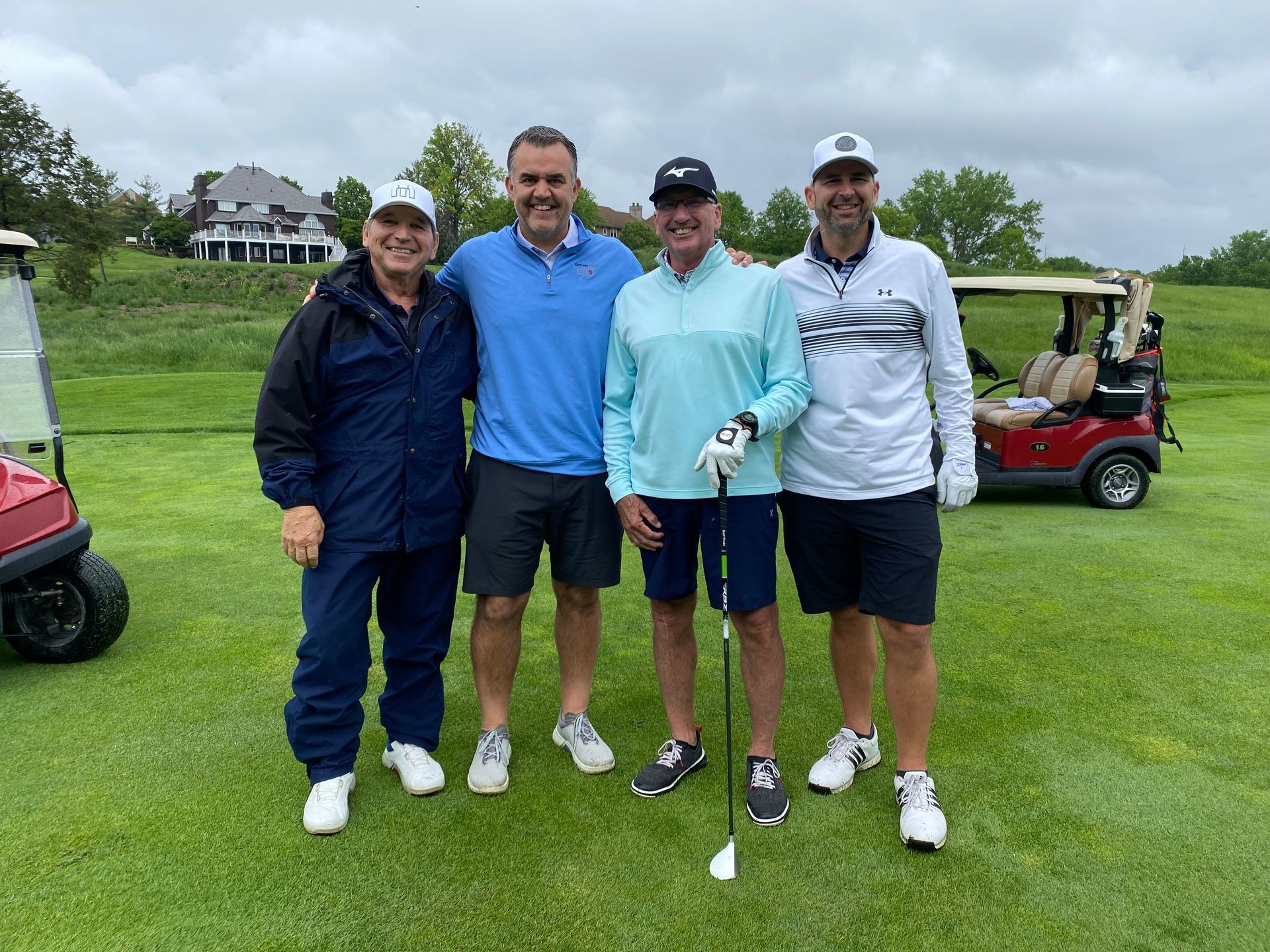 Four men pose on a golf course, smiling in casual golf attire, with golf carts in the background.