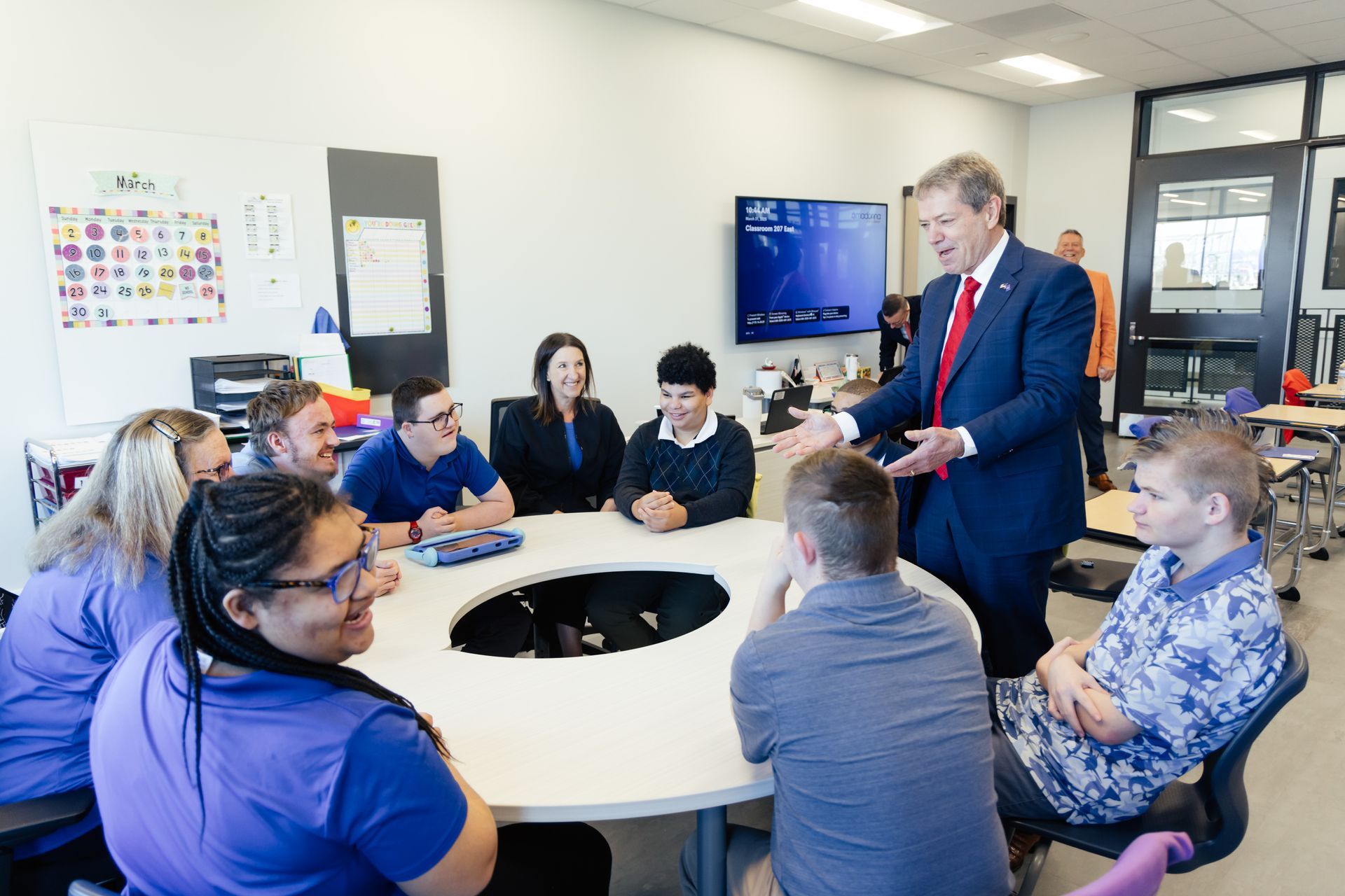 A man in a suit speaks to a group seated around a round table in a classroom.