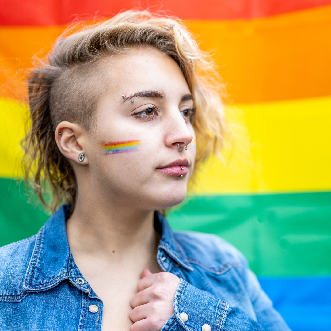 A woman with a rainbow painted on her face is standing in front of a rainbow flag.