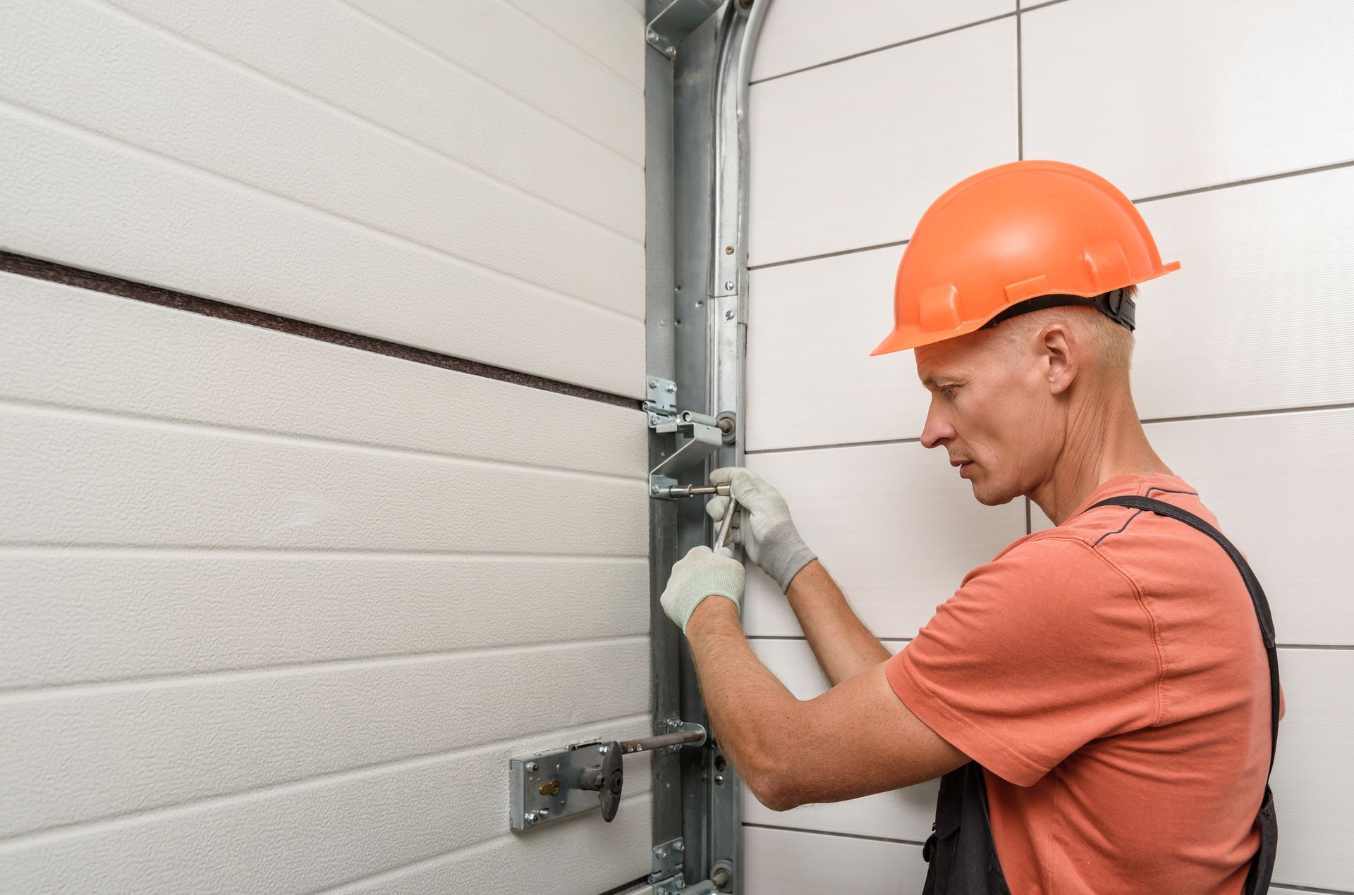 A worker in an orange helmet and gloves repairs a garage door mechanism using tools for maintenance