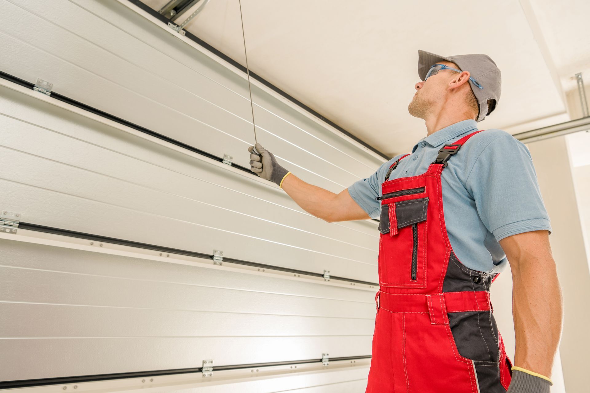 A worker in red overalls adjusts a garage door mechanism with a cable inside a garage