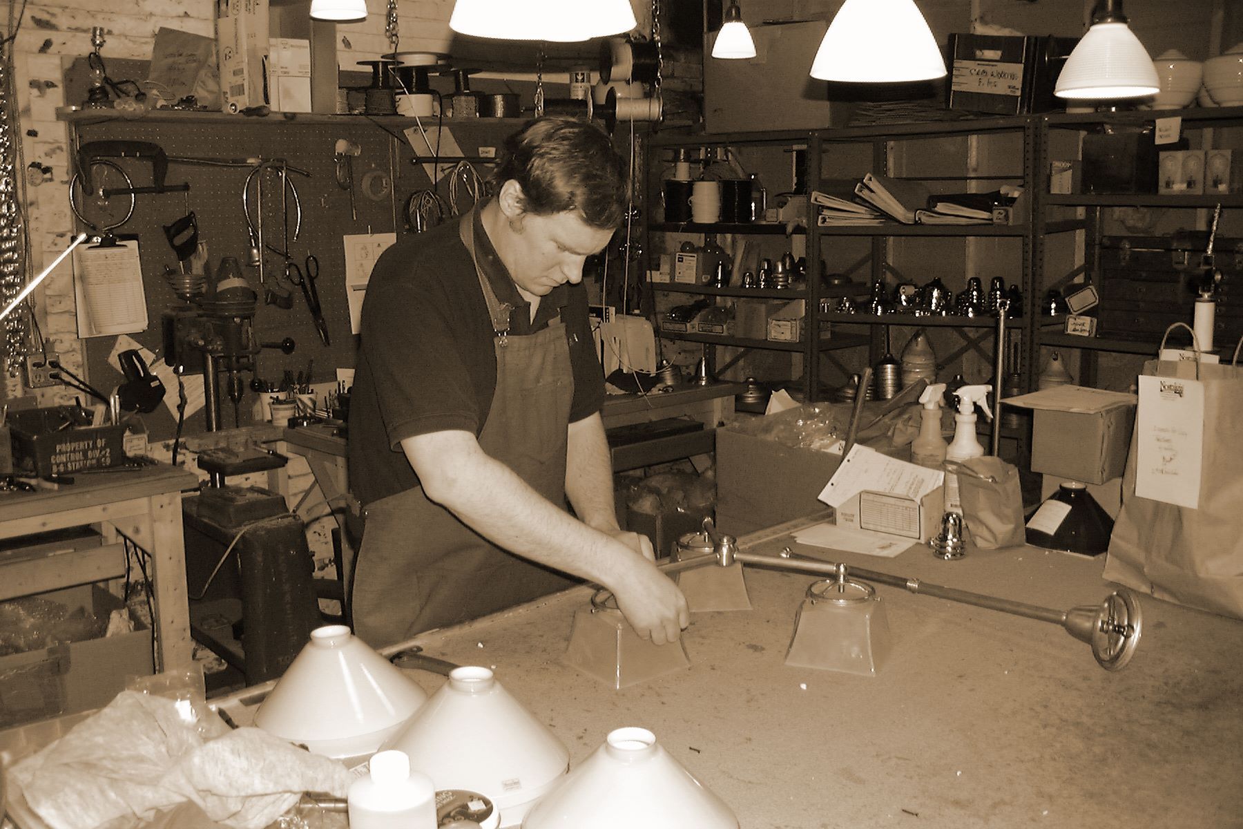 Man working in a workshop with tools, wearing an apron, focused on the task at hand. Sepia-toned.