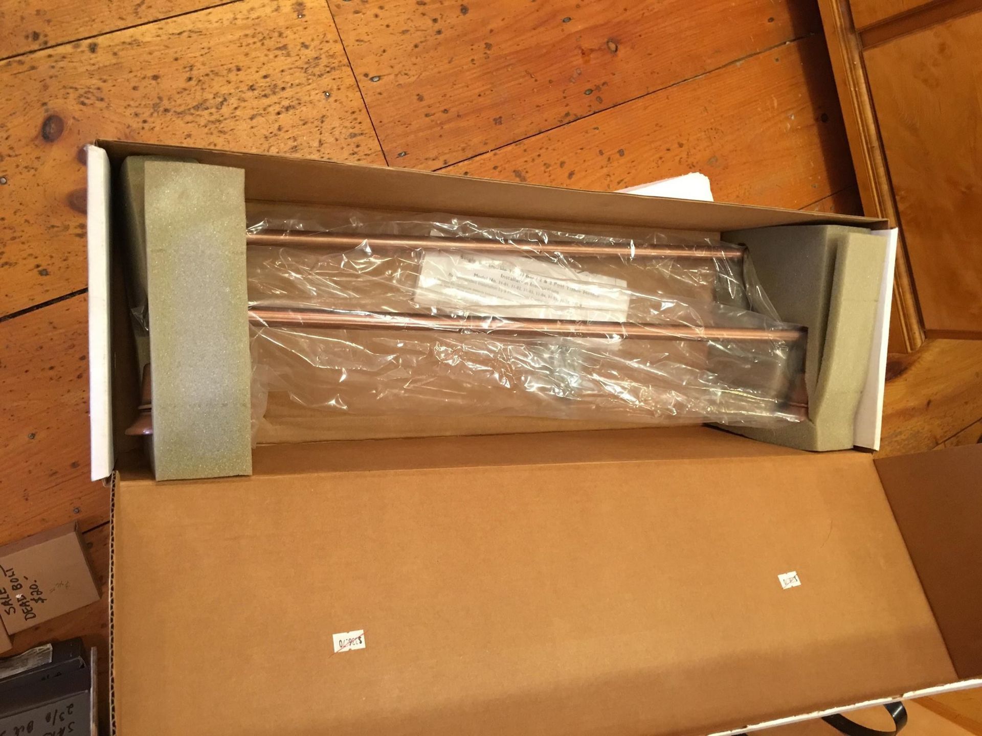 A rectangular box with a copper towel rack wrapped in plastic, sitting on a wooden floor.