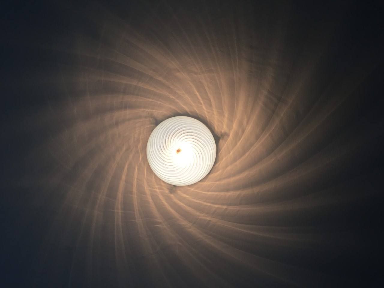 Overhead view of a bright, round light fixture casting a spiraling pattern of shadows on a dark ceiling.