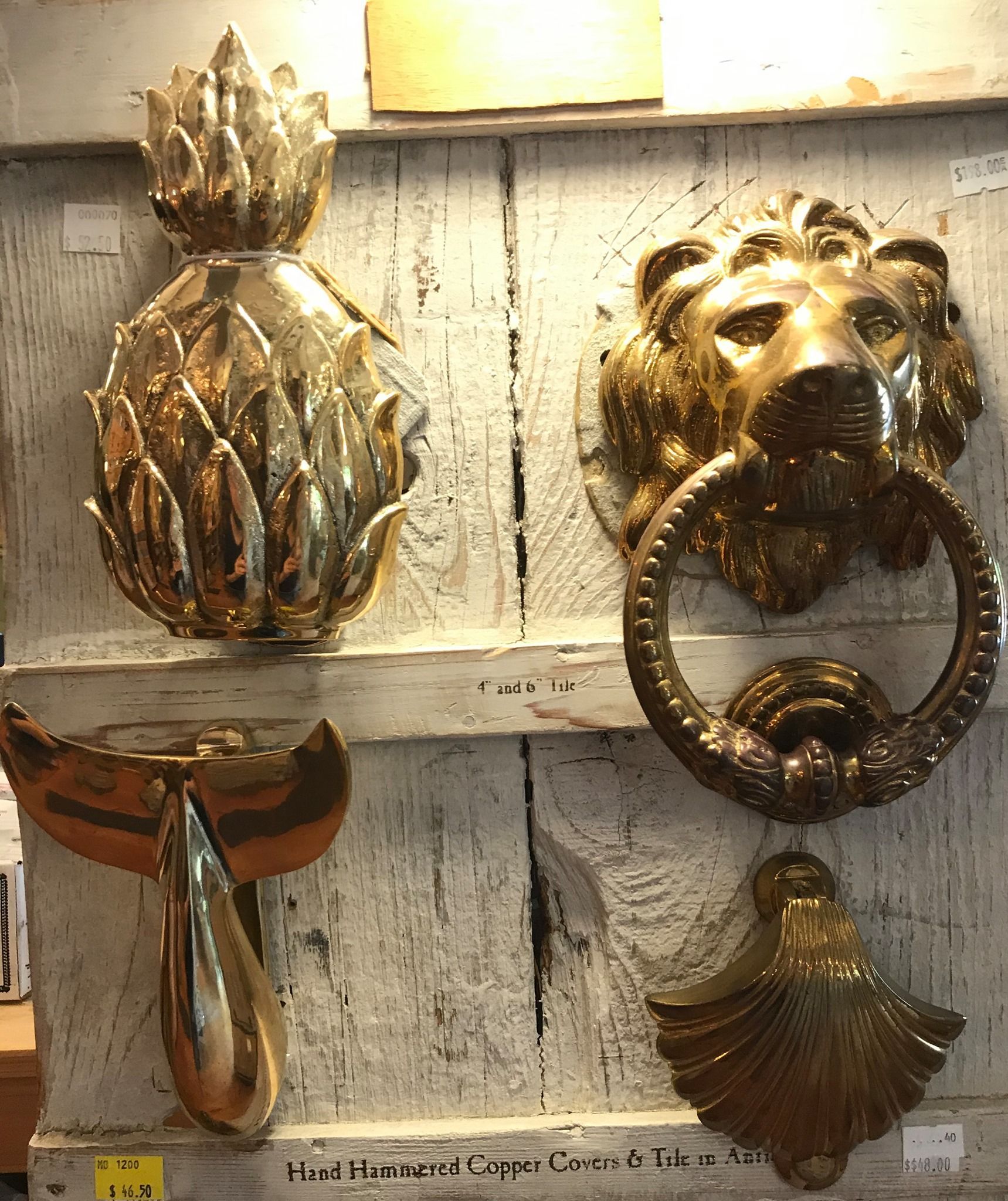 Four ornate gold door knockers on a distressed white surface: pineapple, lion, teardrop, and shell.