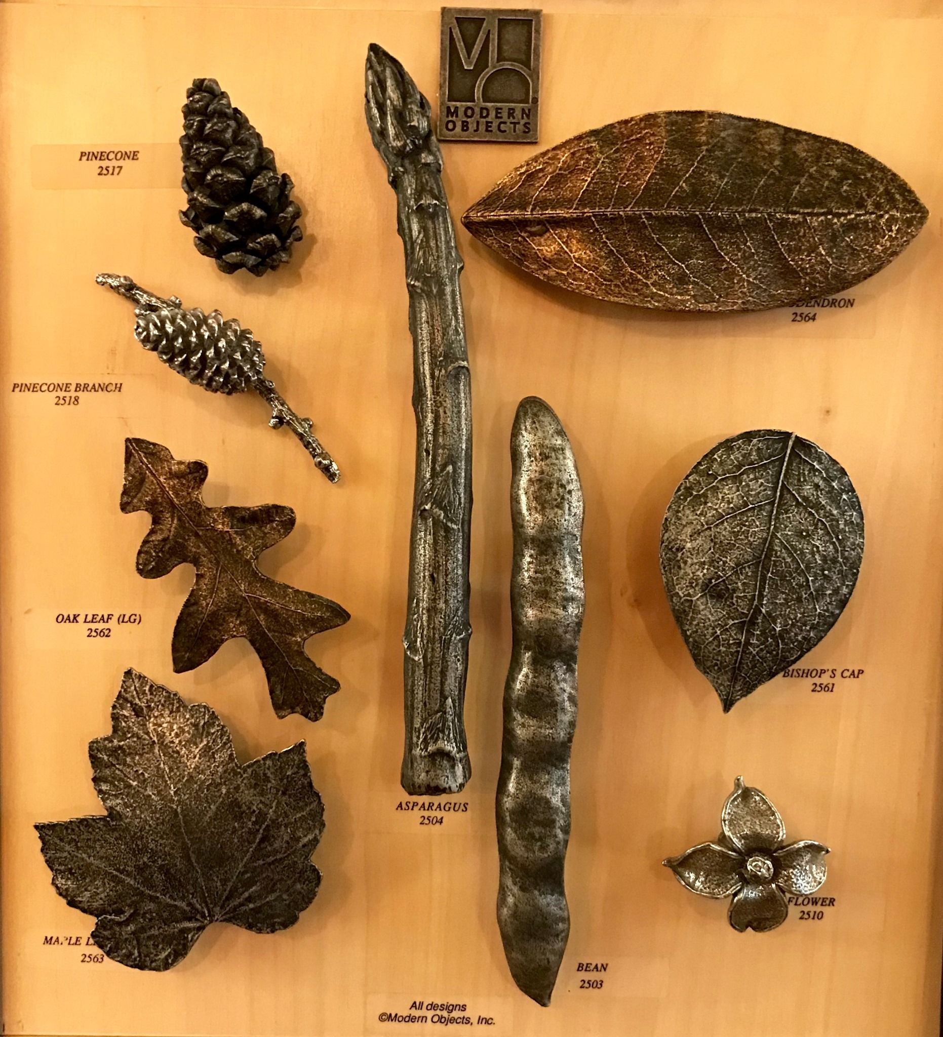Display case of metal sculptures depicting natural forms, including leaves, a pinecone, and asparagus.
