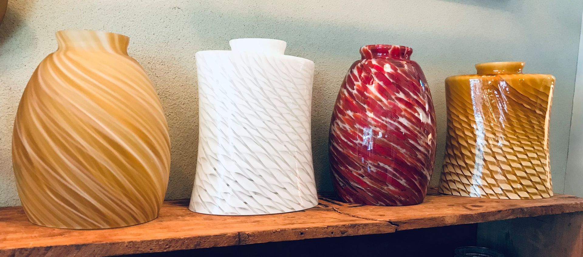 Four colorful glass vases on a wooden shelf. Two are rounded, and two are square, all with swirling patterns.