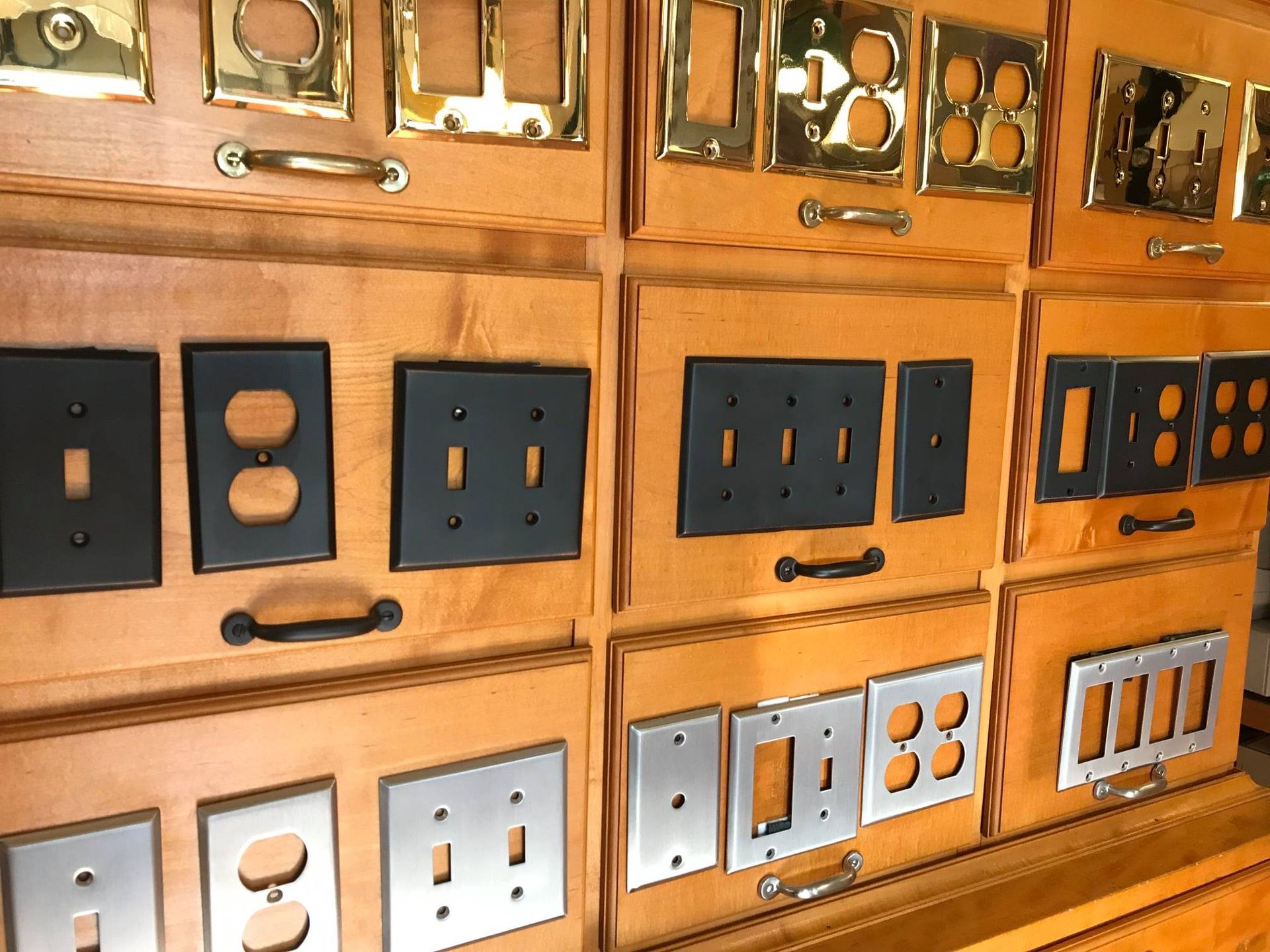 Display of various metal switch plates in brass, black, and silver mounted on a wooden surface.