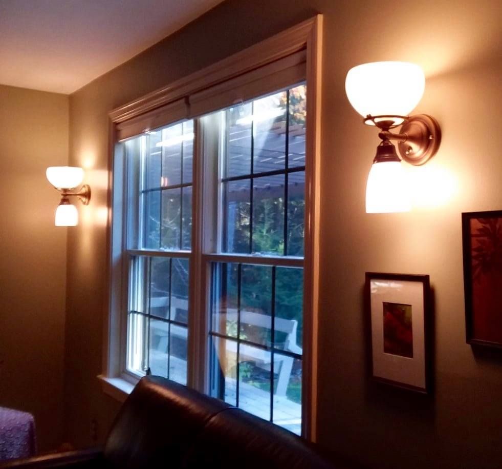 Two wall sconces flank a window in a room with a brown leather couch and artwork.