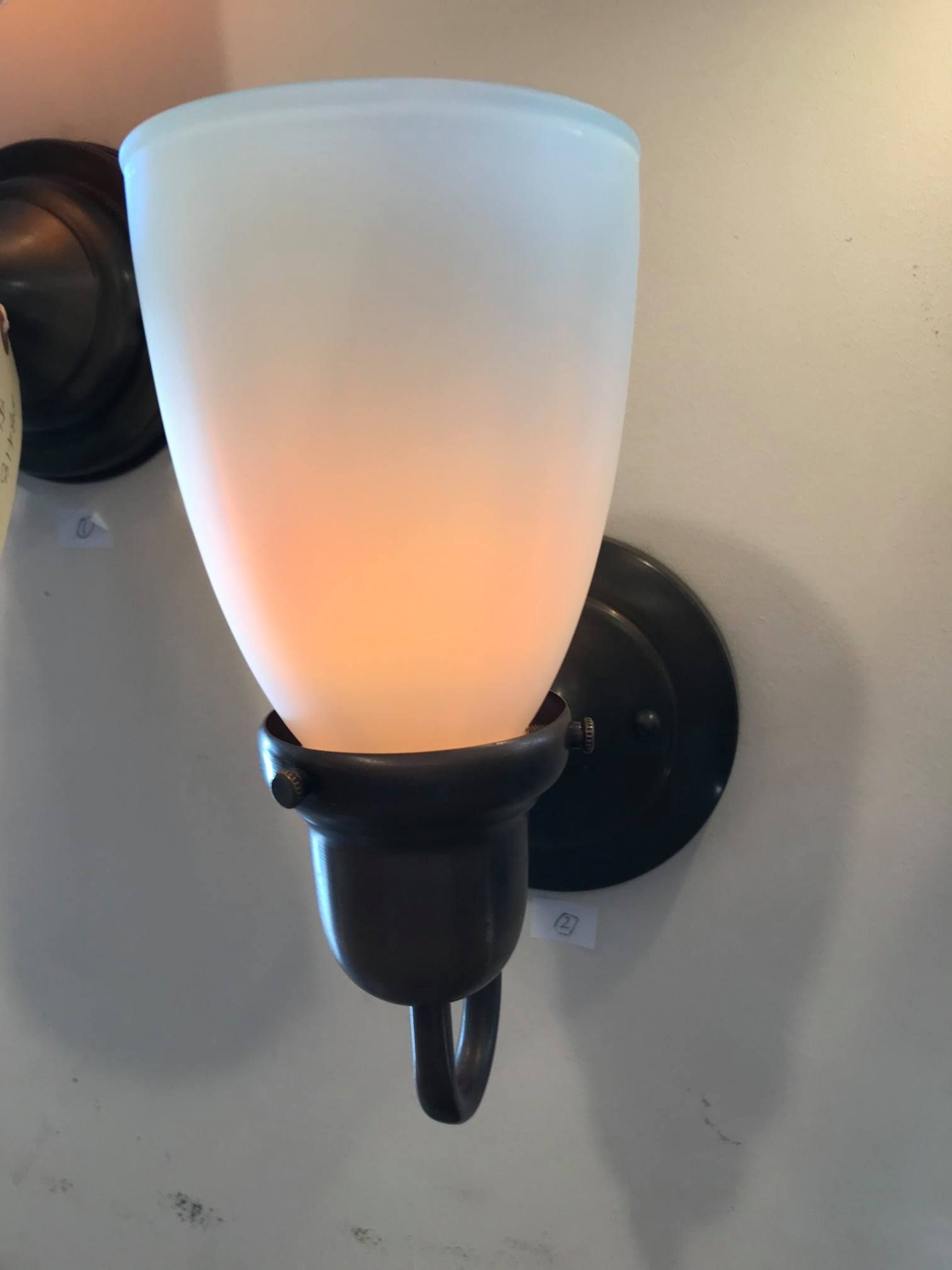 Dark bronze wall sconce with frosted glass shade, illuminated, mounted on a light gray wall.