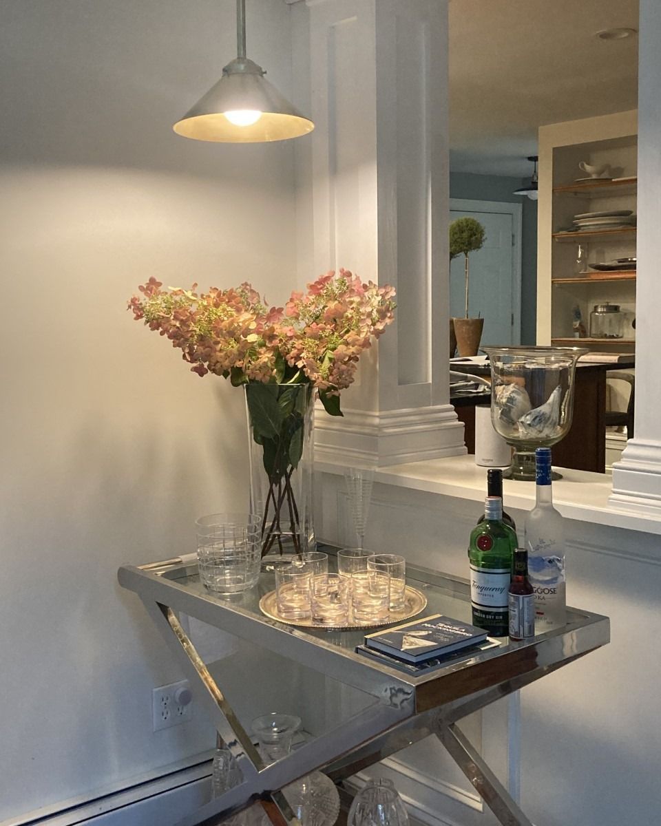 A bar cart with a floral arrangement, glassware, and bottles, illuminated by a pendant light.