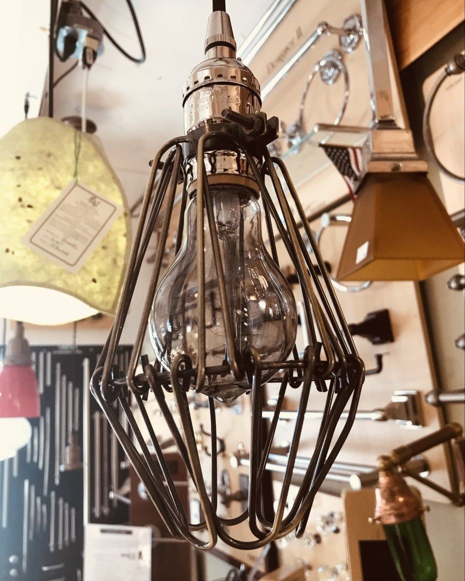 Industrial-style light fixture with metal cage and Edison bulb hanging in a shop, surrounded by other lighting.