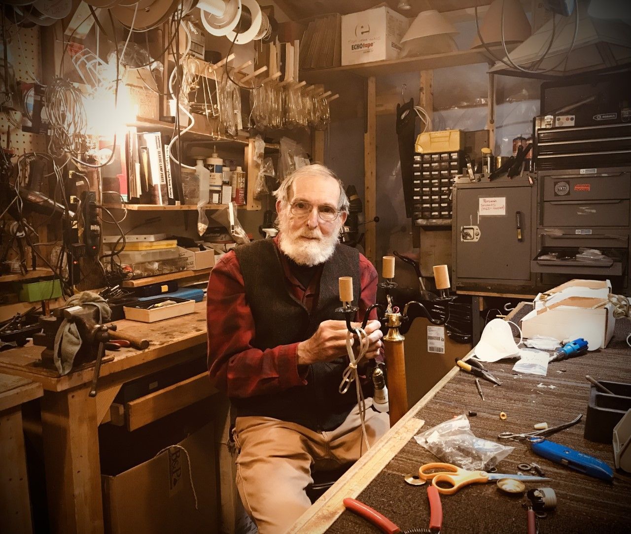 A man with a white beard works on a musical instrument at a workbench in a workshop.