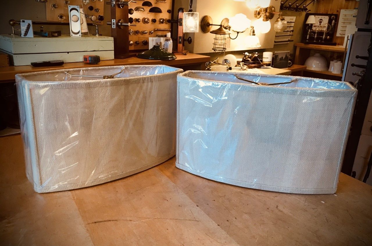 Two oval beige lampshades wrapped in clear plastic on a wooden surface in a store.