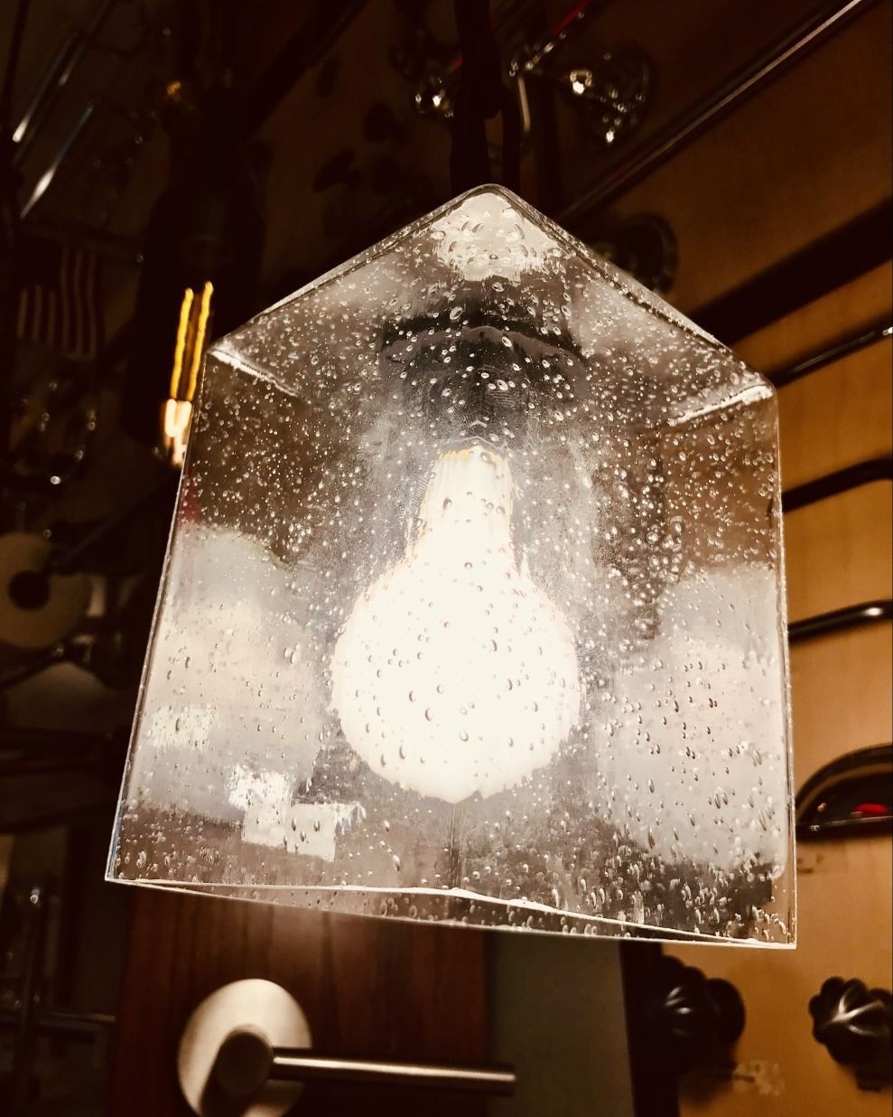 A clear glass, house-shaped lamp with a glowing bulb inside, hanging against a dark background.