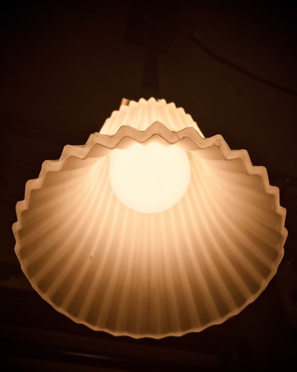White, fluted lampshade with a scalloped edge surrounds a lit bulb, casting a warm glow.