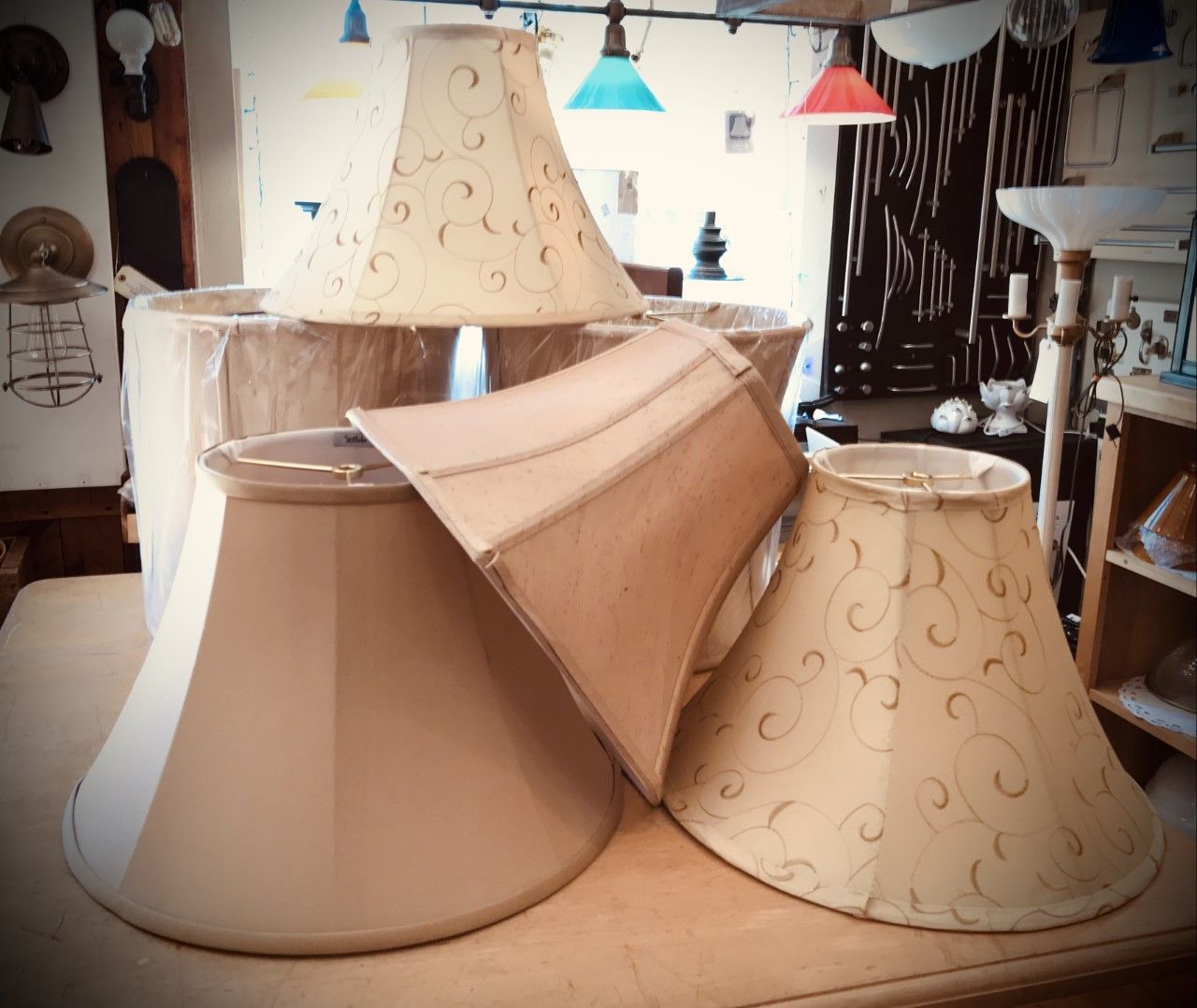 Assortment of beige and tan lampshades, some with scroll patterns, displayed on a wooden surface in a shop setting.