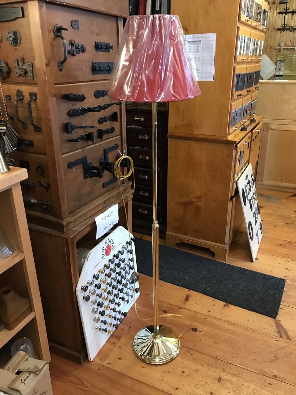 Gold floor lamp with red shade in a shop, next to wooden cabinets and display board.