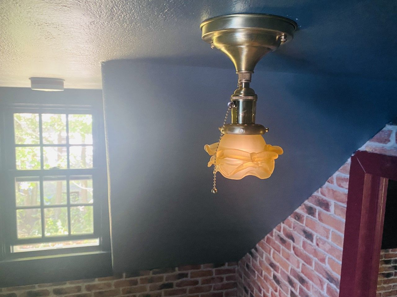 Brass ceiling light fixture with a yellow glass shade, in a room with a window and brick wall.
