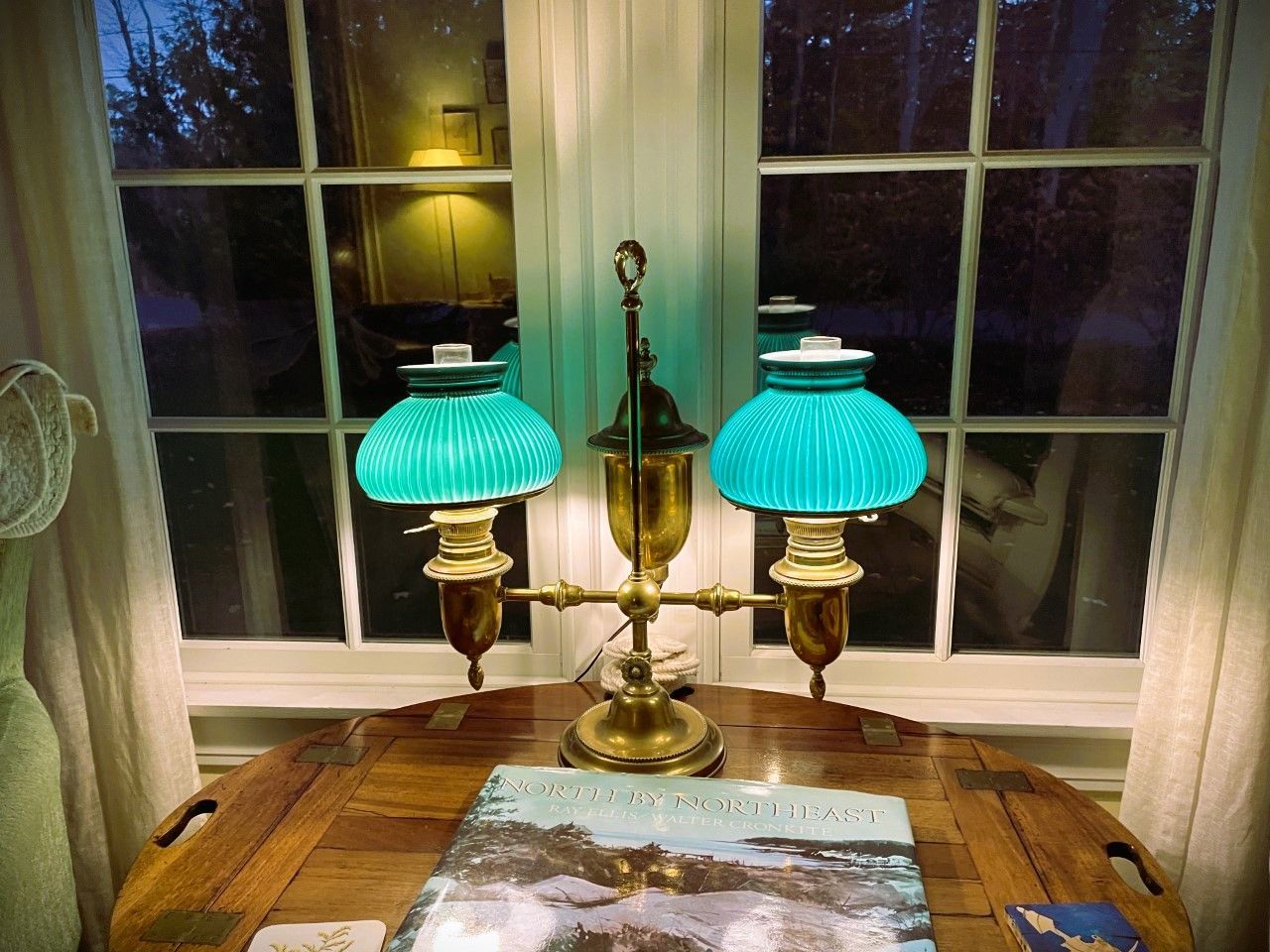 A brass lamp with two turquoise shades sits on a wooden table in front of a window.