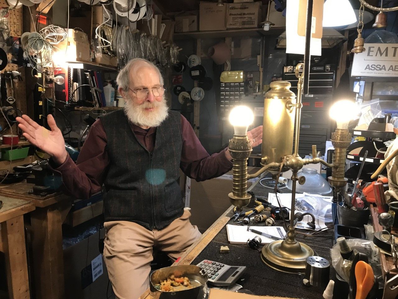 Man with beard, gesturing, seated in workshop with lamp, tools, and clutter.