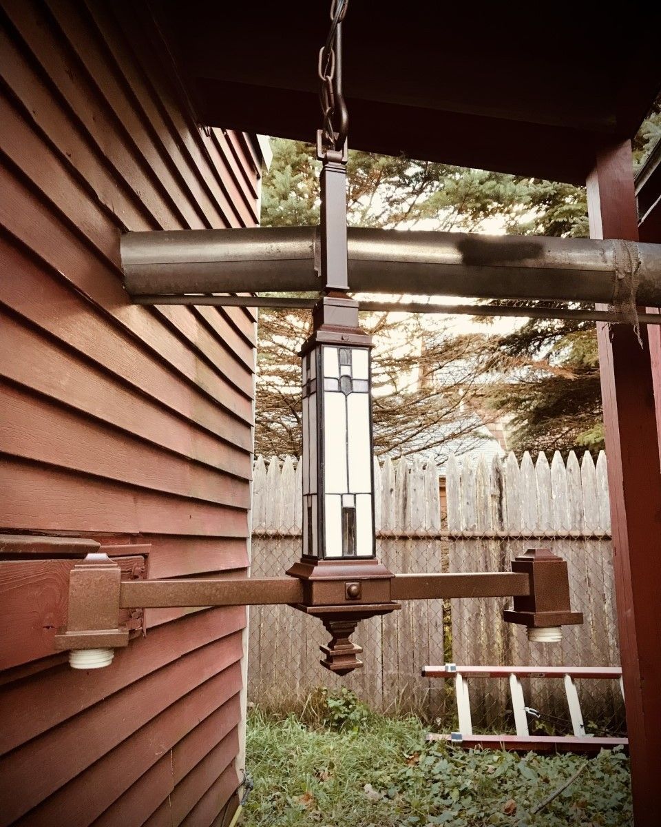 Brown outdoor light fixture hanging on a red building exterior, with a gutter and fence in the background.