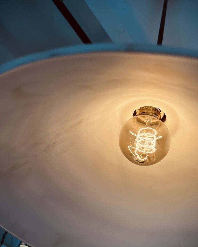 Lightbulb in a white lampshade, glowing with a warm orange light. Wooden beams visible above.