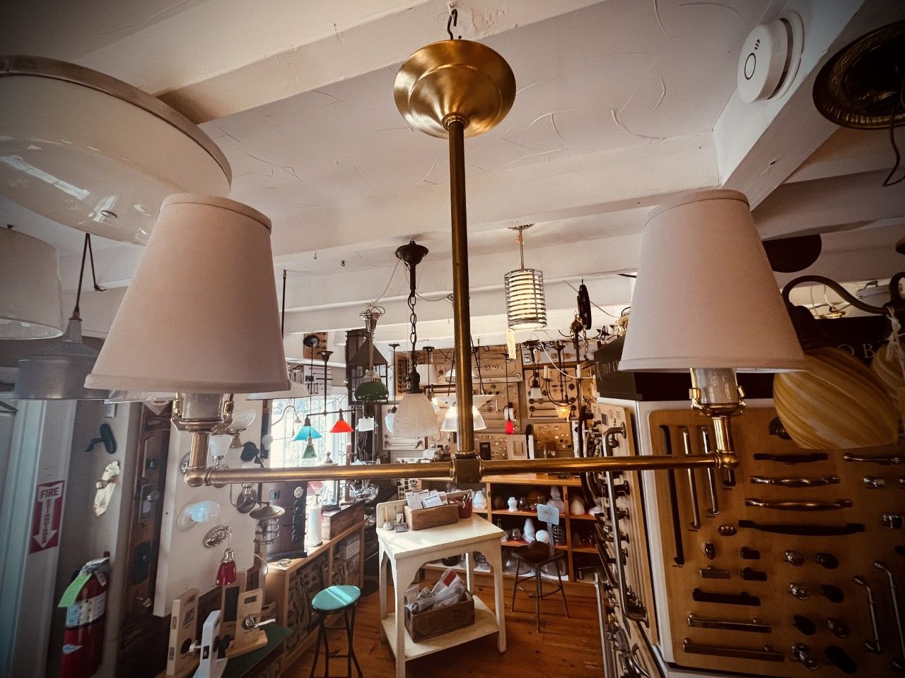 Brass chandelier with two lampshades hanging in a shop, surrounded by items for sale.