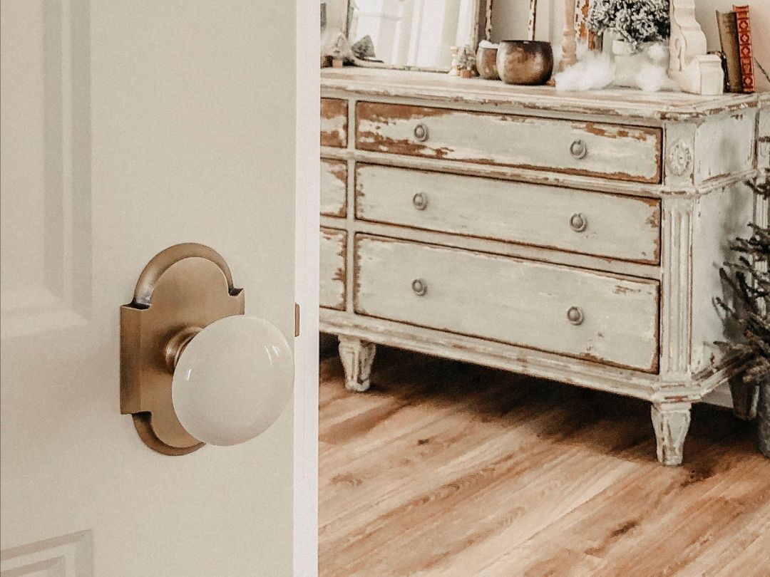 White door with brass hardware partially open, revealing a distressed white dresser on wood flooring.