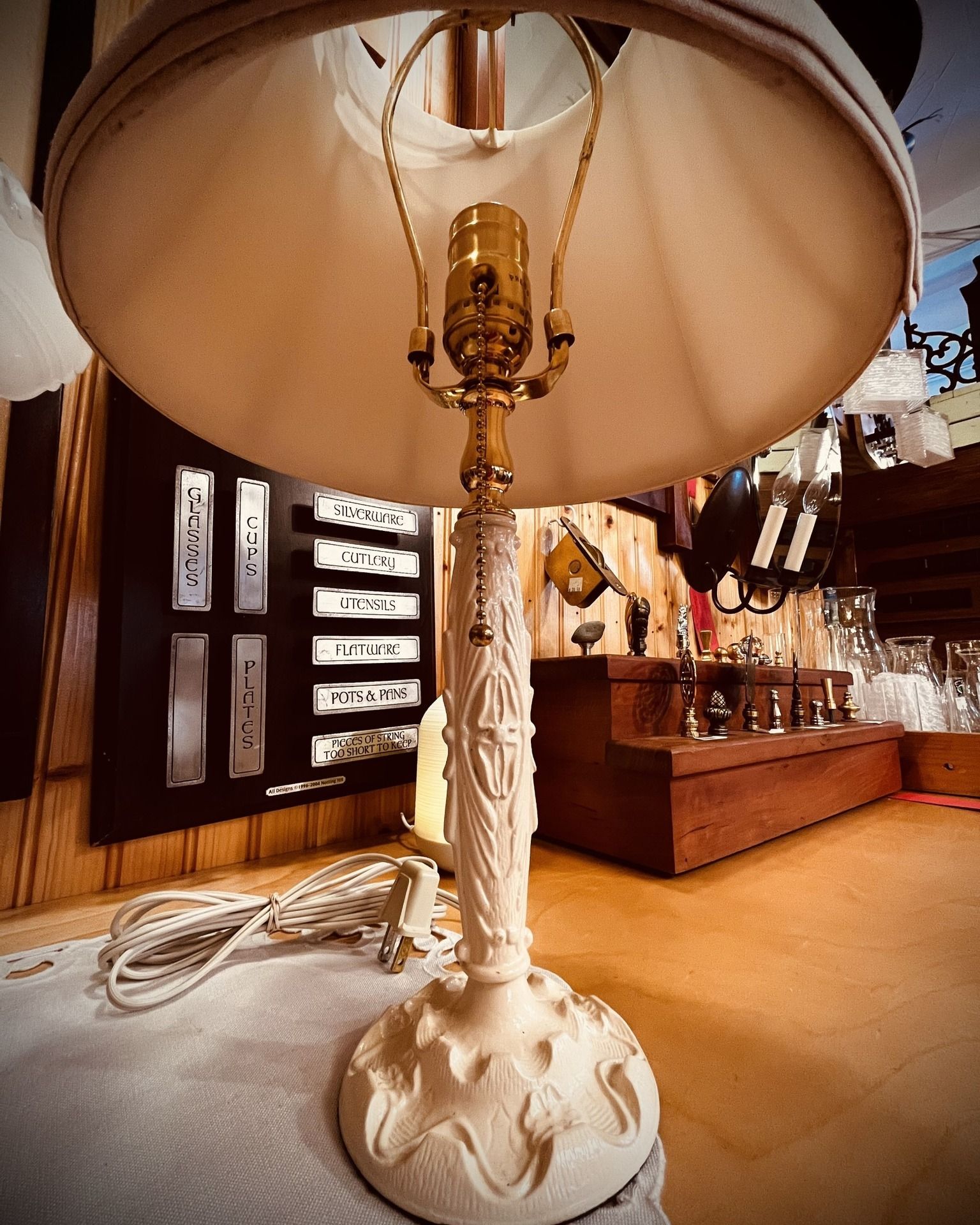 Cream-colored antique lamp with carved base and shade on a wooden surface with a wall display in the background.