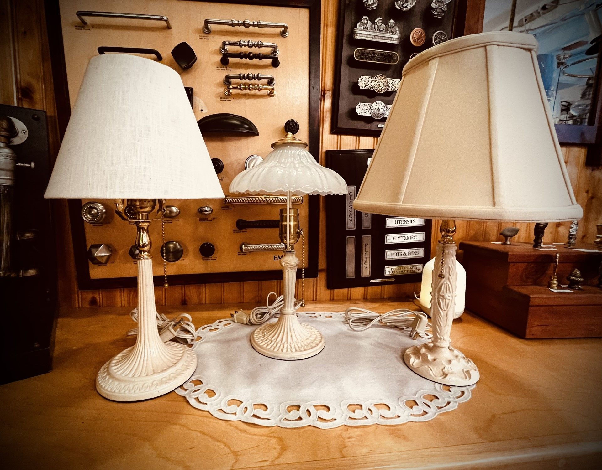 Three ornate cream-colored lamps with lampshades on a lace doily, set against a wall of hardware.