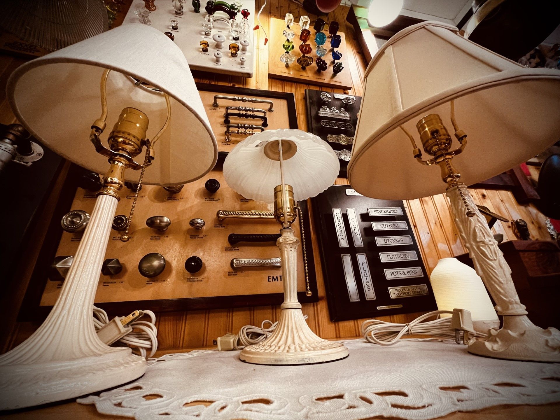 Three antique lamps with shades on display in a shop, with hardware on the wall behind them.