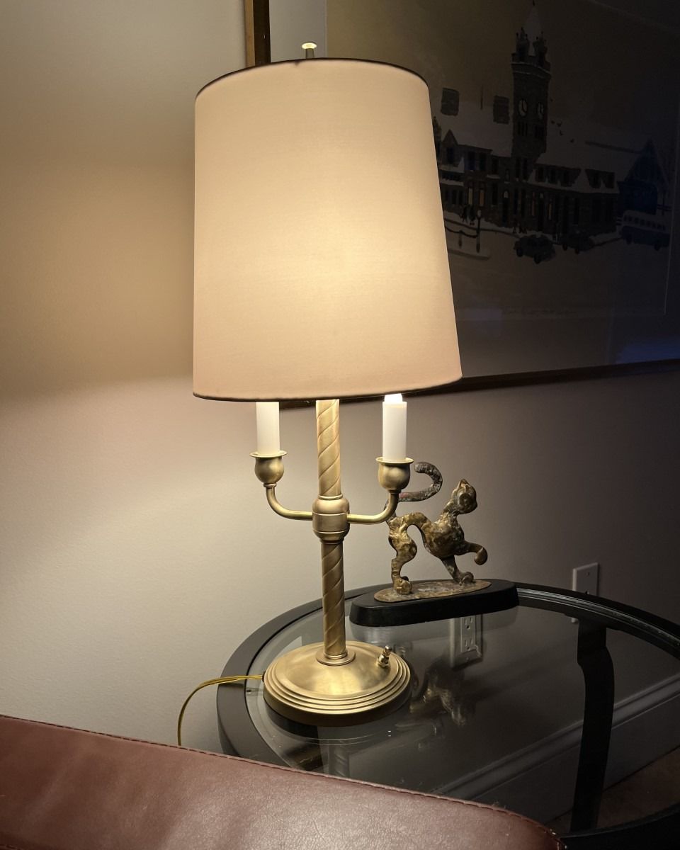 Brass lamp with a tan shade on a glass table next to a decorative figurine.